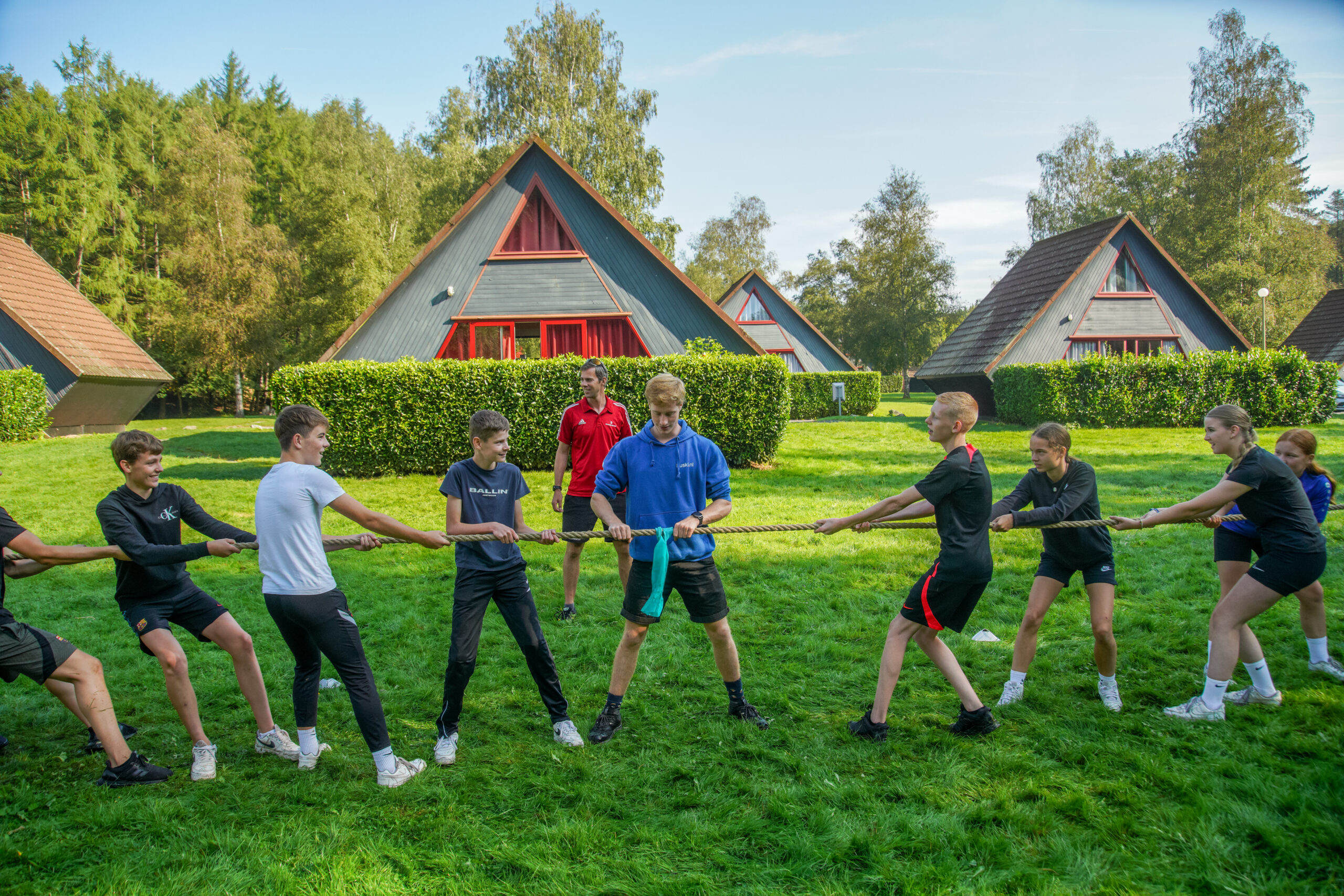 Een groep jongeren speelt touwtrekken op een grasveld tijdens een zomerkamp, met vakantiehuisjes en bomen op de achtergrond.