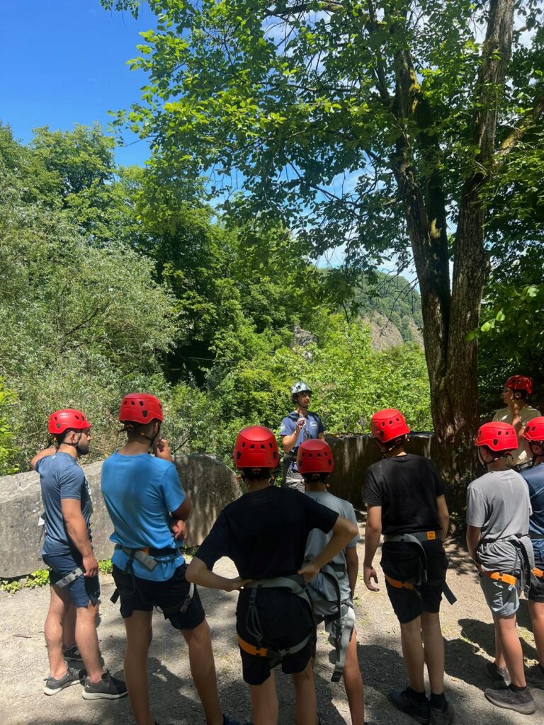 Een groep jongeren met rode helmen en klimgordels luistert buiten naar instructies van een begeleider in een bosrijke omgeving.
