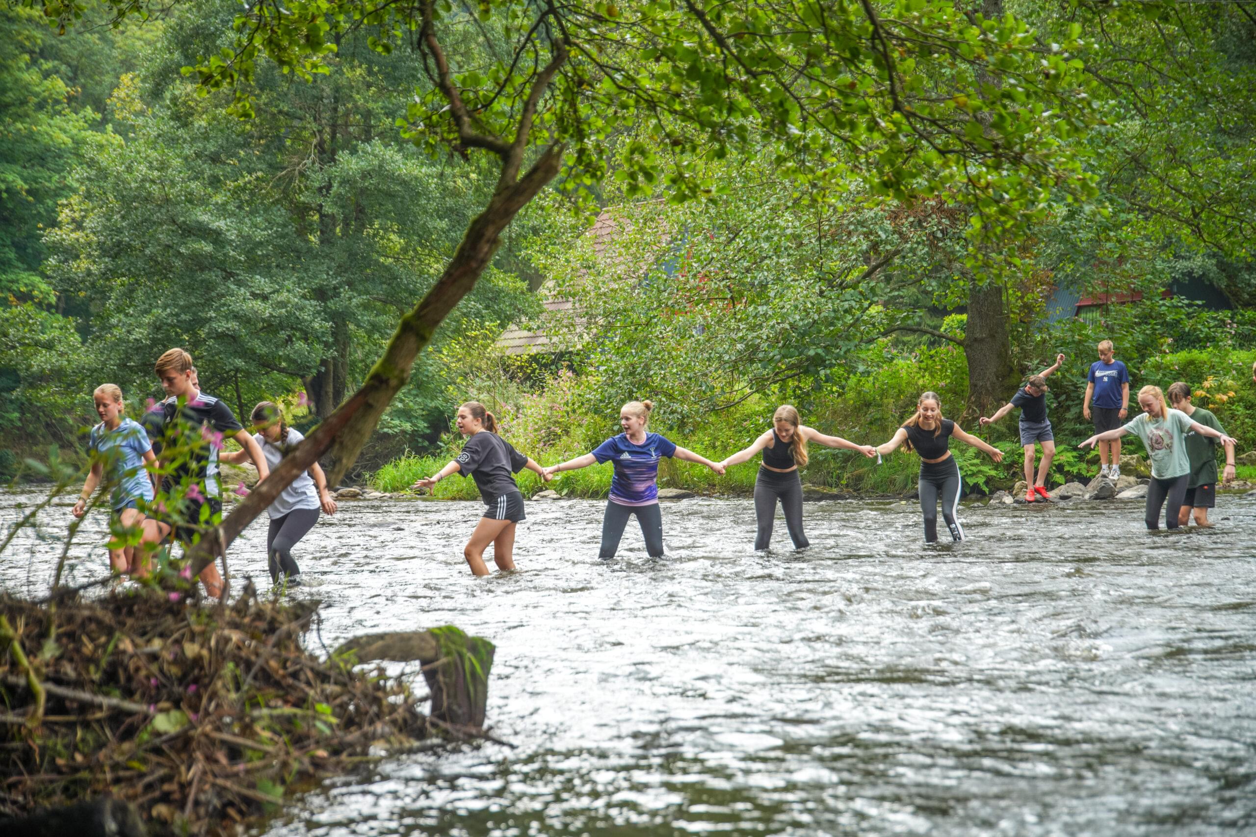 Een groep jongeren loopt hand in hand door een ondiepe rivier, omringd door een groene, bosrijke omgeving.